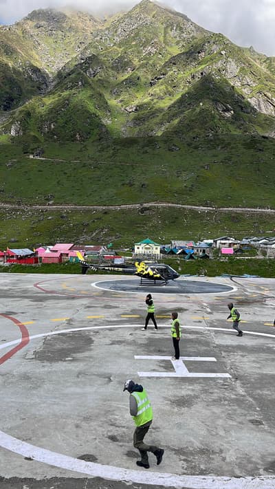 Ground crew in high-visibility vests on the concrete helipad at Kedarnath as a small helicopter sits on the marked pad, with the village and a green mountainside behind