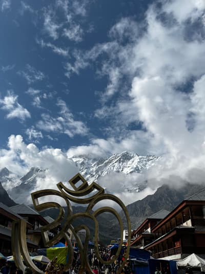 A large golden Om sculpture standing over the market street at Kedarnath, with timber-fronted shops either side and snow peaks above the valley head