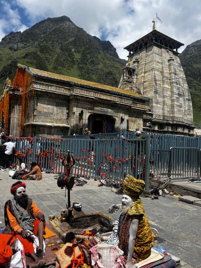 Two ash-covered sadhus seated beside a small fire in front of Kedarnath temple, with pilgrims' shoes left at the railing and red threads tied along the fence