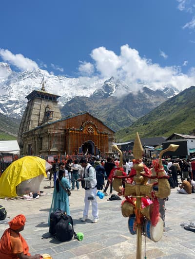 Kedarnath temple with its facade draped in marigold garlands, snow-covered peaks of the Kedarnath massif rising behind, and pilgrims gathered across the stone forecourt
