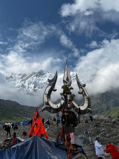 A large polished steel trident hung with rudraksha beads standing on the open moraine at Kedarnath, with snow-covered peaks emerging from cloud behind