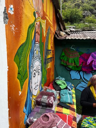 Hand-knitted sweaters, socks and gloves laid out on a checked cloth at a roadside stall in Mana village, beside a brightly painted wall mural